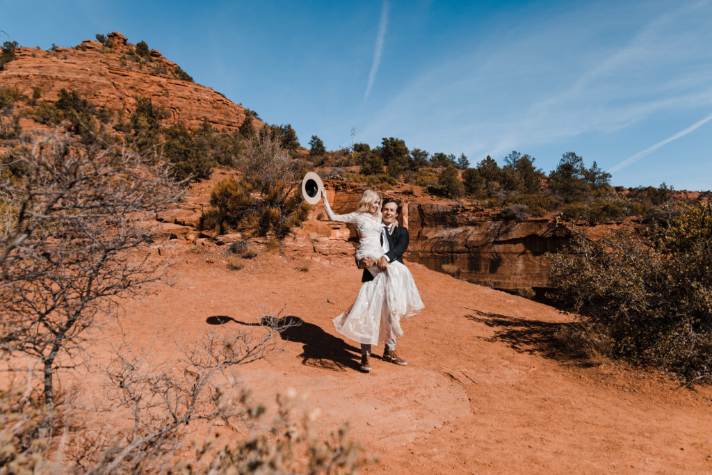 A couple in wedding attire in Sedona Arizona for their elopement. The groom is carrying the bride in his arms while the bride waves her hat.