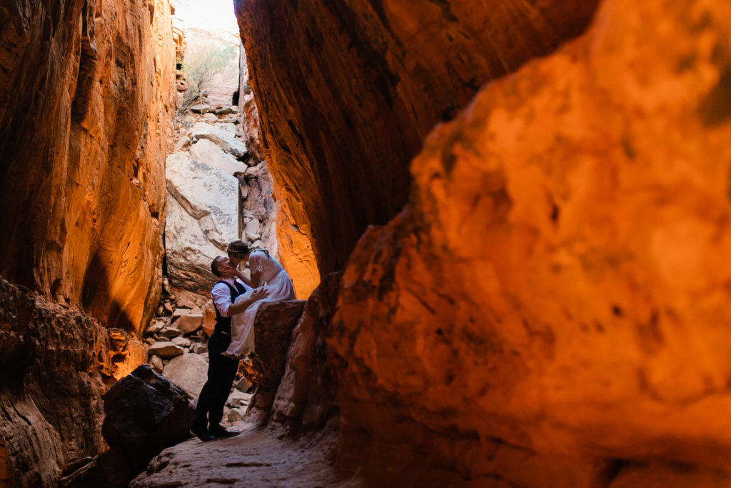 A couple in wedding attire in a red rock cave. One person is wearing a white dress and sitting on a ledge while the other person in a suit faces and kisses them. 