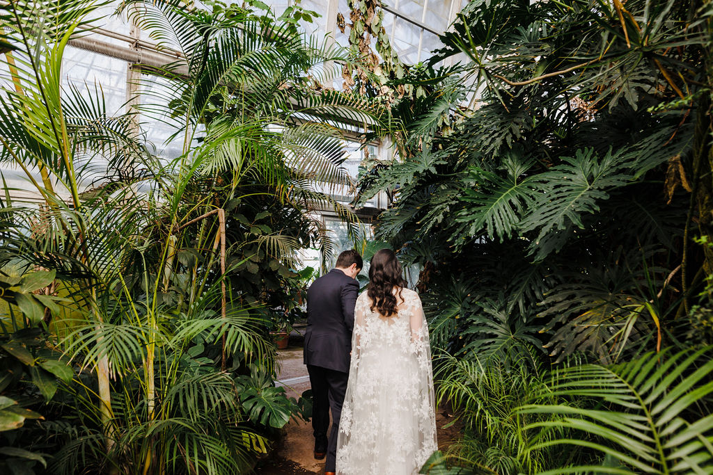 A couple in wedding attire walks through ferns in a greenhouse