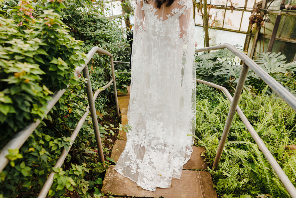 A close up of a wedding dress train as a bride walks down stairs surrounded by greenery