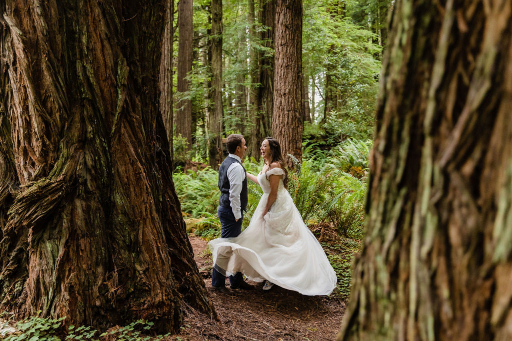 A couple in wedding attire twirls in the woods in between tall redwood trees 
