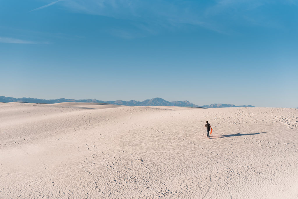 A person walking alone in the white sand dunes, holding an orange disc sled.